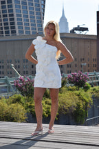A woman stands outdoors on a rooftop terrace wearing a white one-shoulder mini dress with large sculptural flower-like appliqués and clear strap heels, with city buildings and greenery in the background.
