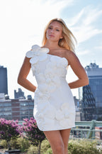 A blonde woman wearing a white one-shoulder mini dress with large 3D flower appliqués, posing outdoors against a city skyline background.