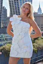 A blonde woman wearing a white one-shoulder mini dress with large 3D floral appliqués, posing outdoors with a city skyline background.