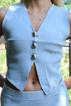 A close-up of a person wearing a light blue ribbed knit sleeveless top with a deep V-neck, three large metallic buttons down the front, and a cutout at the waist, paired with a matching skirt.
