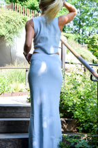 A woman wearing a light blue sleeveless dress with side cutouts, photographed from behind on outdoor wooden steps with greenery and a metal railing in the background.