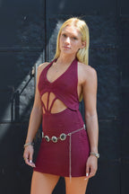 A blonde woman wearing a maroon ribbed halter mini dress with front cutouts and a silver chain belt, standing against a dark metal door.